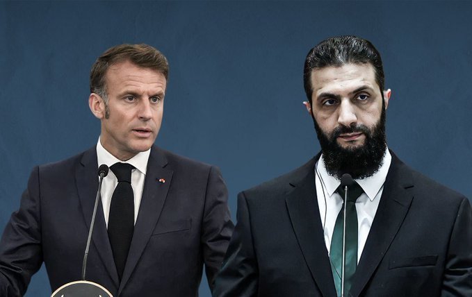 Two suited men converse face-to-face while a third bearded man stands between them, with several national flags visible in the blurred background.