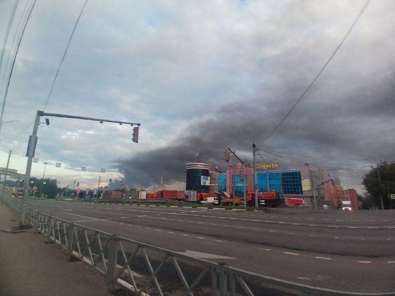 A plume of dark smoke rises above a commercial building and a striped tower beyond an empty multi-lane road.