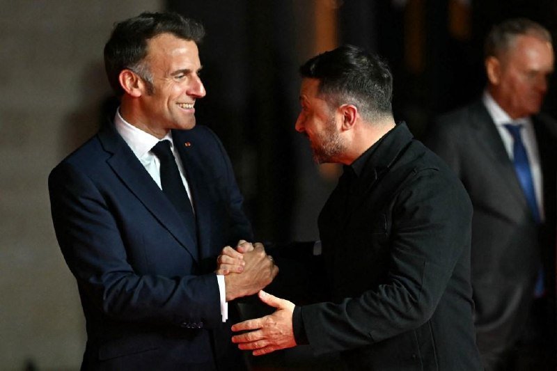 Two men shake hands in front of American and Ukrainian flags, one in a black shirt and the other in a dark suit with a purple tie.