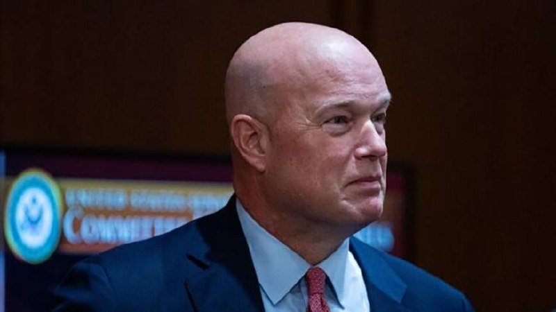 A bald man in a dark suit, light blue shirt, and red patterned tie stands in front of a screen displaying a government seal and partial text.