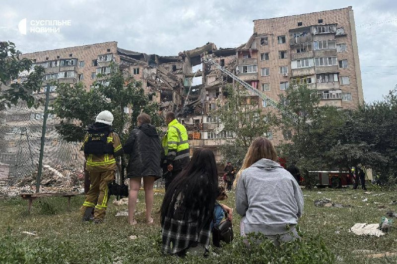 Firefighters in uniform and civilians observe a heavily damaged multi-story residential building with collapsed sections and shattered windows.