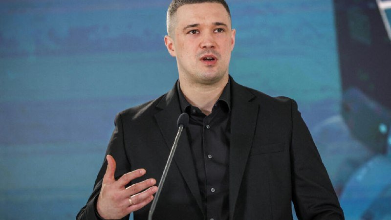 A man in a black suit and dark shirt speaks at a microphone against a blue background.