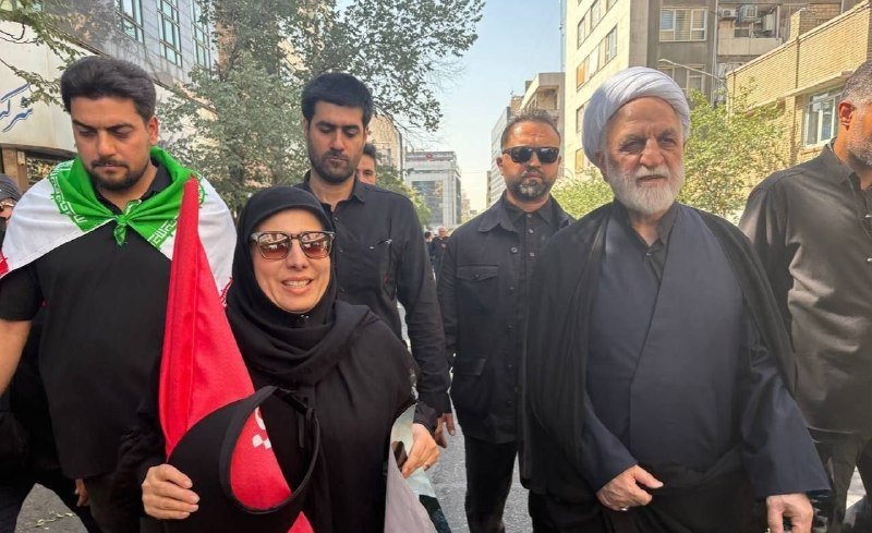 A group of people, including a woman in a black chador and a cleric in traditional robes, walk down a city street, one man draped in an Iranian flag.