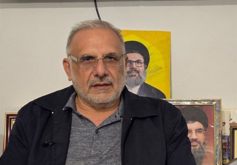 A gray-bearded man wearing glasses and a dark jacket speaks indoors, with two framed portraits of bearded men in clerical attire displayed on the wall behind him.