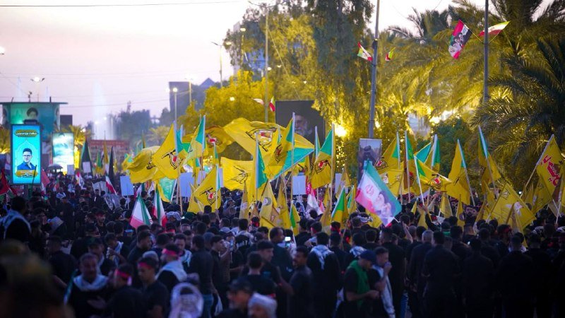 A large crowd marches down a palm-lined street at dusk, waving yellow flags and holding up portraits as banners and posters line the roadway.