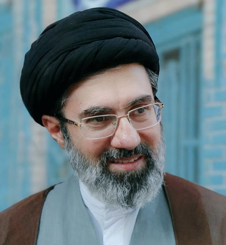 A smiling bearded man wearing a black turban, rimless glasses, and clerical robes stands in front of a blue tiled wall.