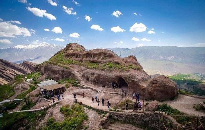 Tourists gather on a rocky mountain overlook featuring a cave-like rock formation, a small pavilion, and distant snow-capped peaks under a partly cloudy sky.