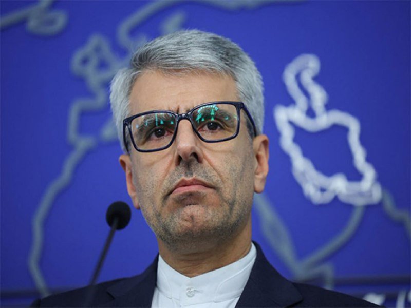 A gray-haired man wearing glasses and a dark suit stands before a microphone against a blue backdrop displaying a white map outline.