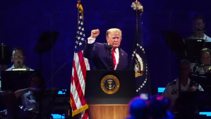 A hand wearing an American flag-patterned sleeve reaches down to press a brass service bell labeled "UNITED NATIONS" against a blue UN emblem backdrop.