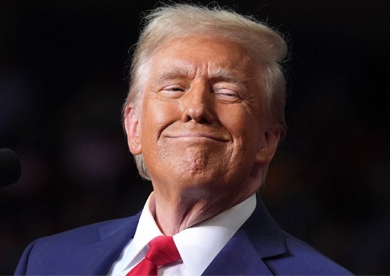 A man with blond hair wearing a blue suit, white shirt, and red tie stands before a dark background with a microphone visible, smiling with eyes closed.