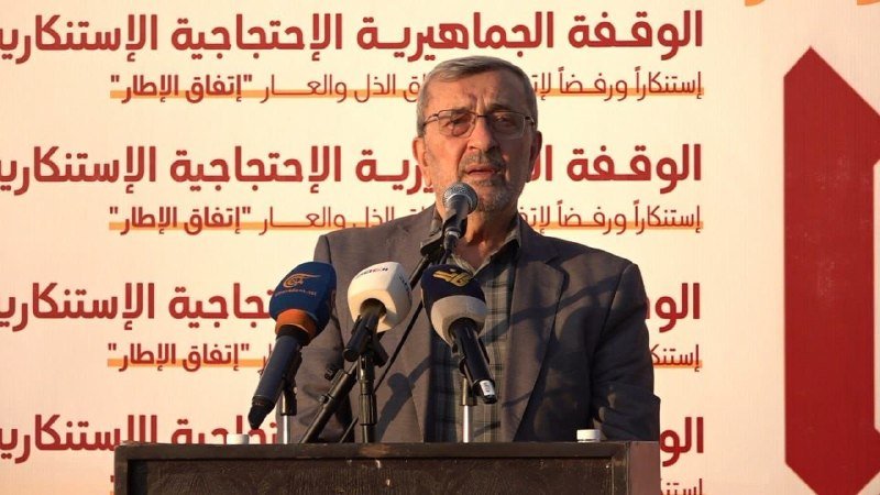 A bearded man in a dark blazer and glasses stands at a podium speaking into multiple microphones, with a backdrop displaying repeating Arabic text.