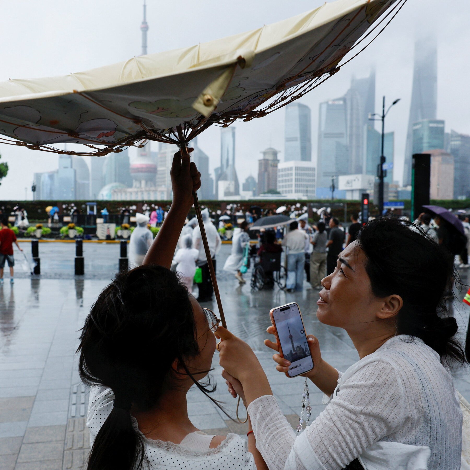 Two women, one holding an umbrella, use a smartphone to photograph a hazy city skyline featuring a tall tower and skyscrapers across a wet plaza crowded with pedestrians.