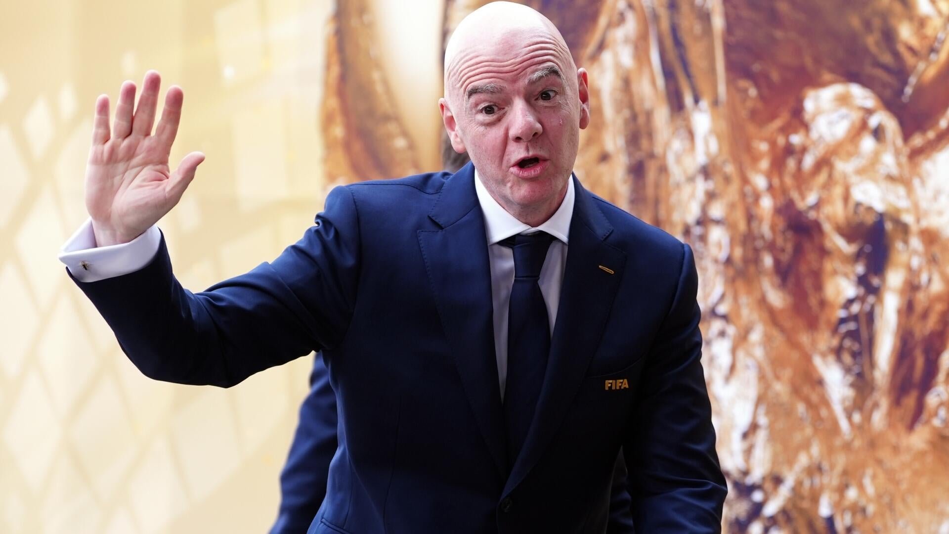 A bald man in a navy blue suit with a "FIFA" lapel pin raises his hand while speaking, standing before a textured golden-brown backdrop.