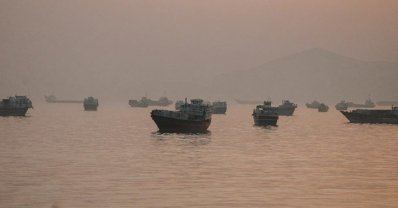 Multiple cargo ships and boats sit anchored in hazy, calm waters with a faint mountain silhouette visible in the background at dusk.