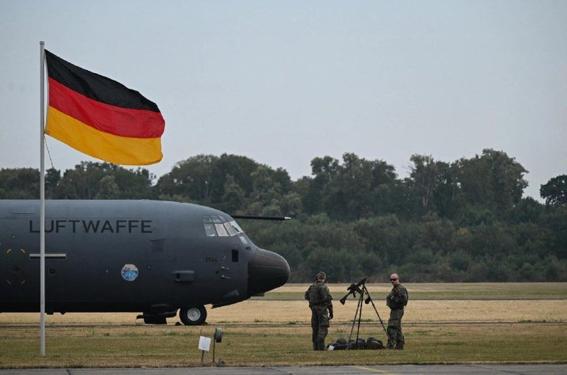 A German flag flies beside a gray military aircraft labeled "LUFTWWA," with two camouflaged personnel standing near a tripod on a grassy airfield.