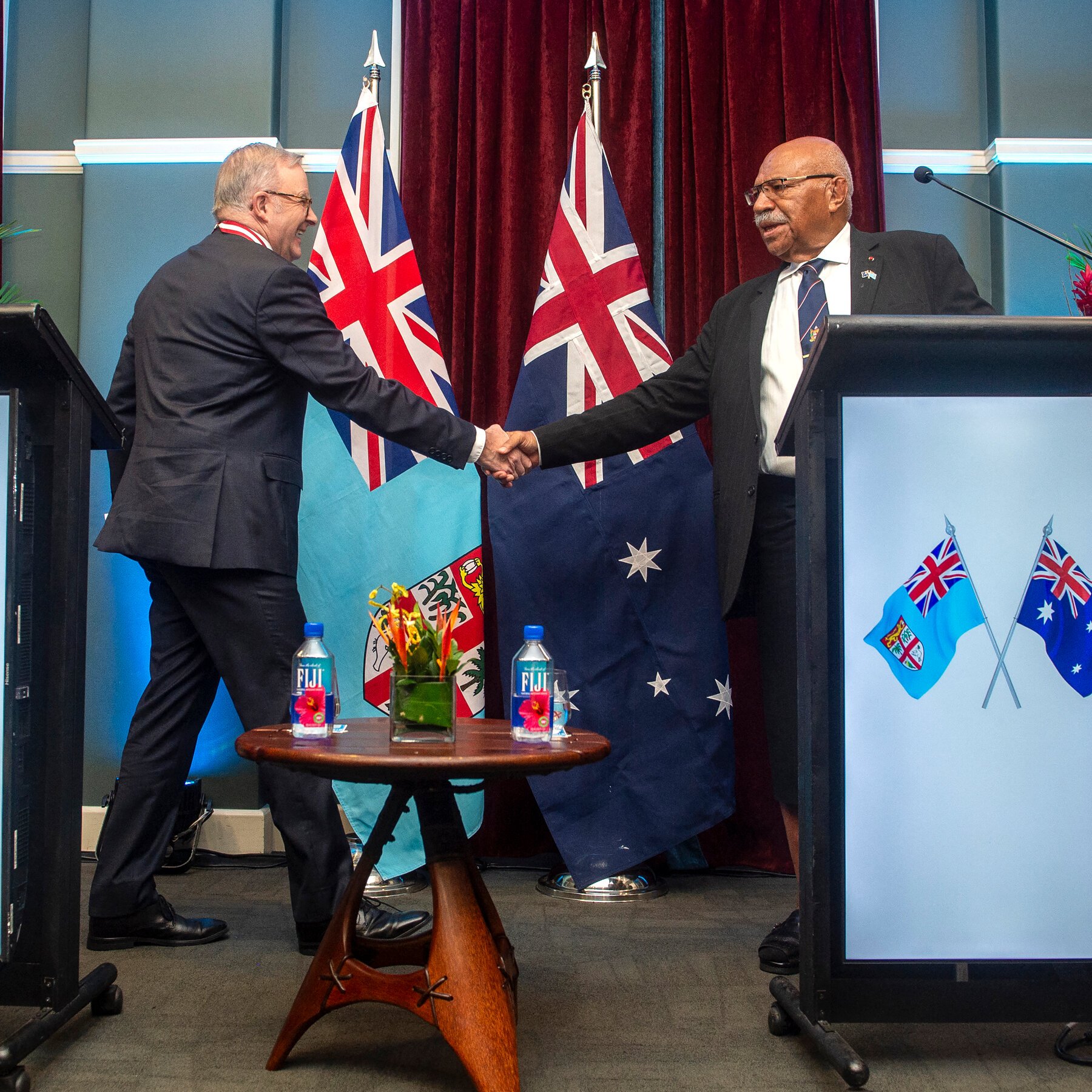 Two men in suits shake hands on a stage flanked by Fijian and Australian flags, with a small table holding Fiji water bottles and flowers between them.