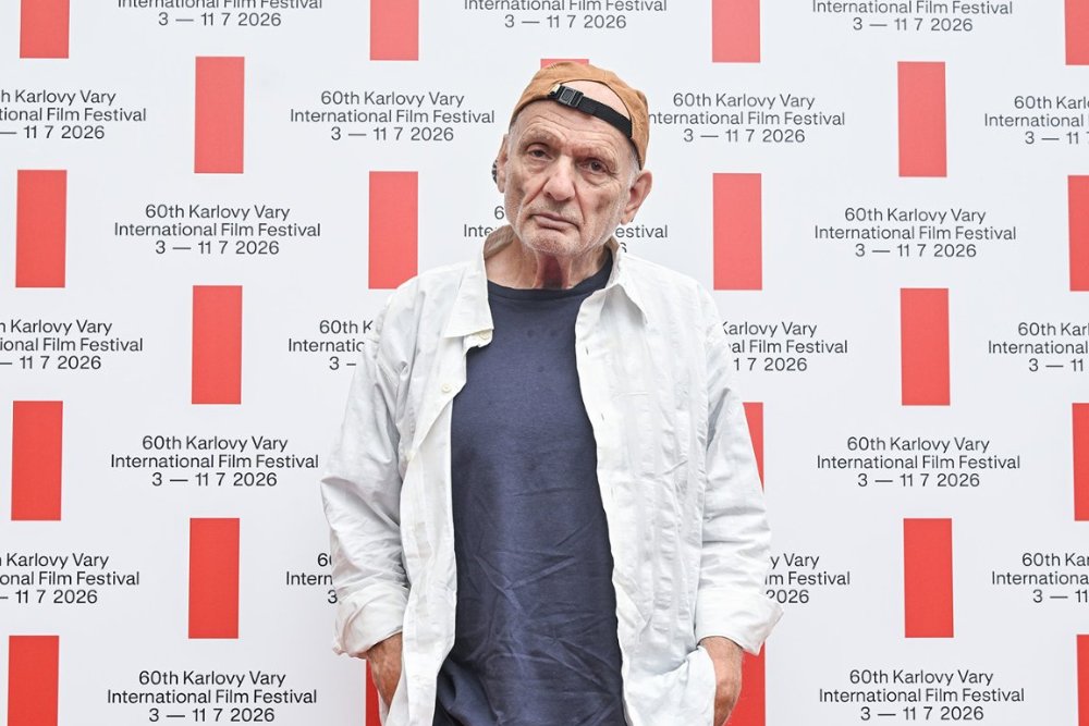 A man wearing a backwards tan cap, white shirt, and blue t-shirt stands in front of a step-and-repeat backdrop reading "60th Karlovy Vary International Film Festival, 3 — 11 7 2026."