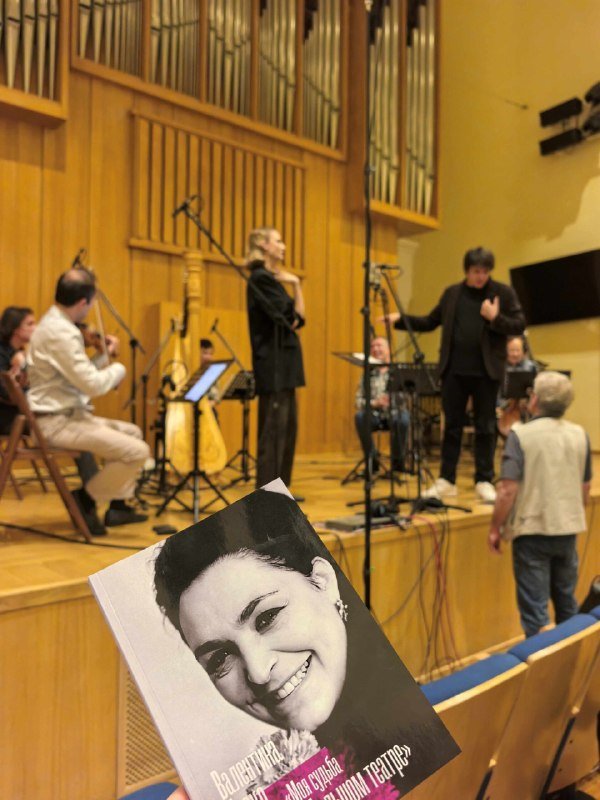 A black-and-white concert program featuring a woman's portrait is held in the foreground, while musicians, singers, and a conductor perform on a wooden concert hall stage.