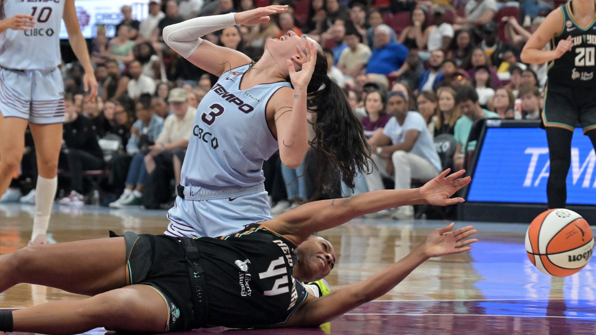 Two WNBA players, one in a white jersey (#3) and one in black (#14), dive for a loose ball on the court as a crowd watches from the stands.