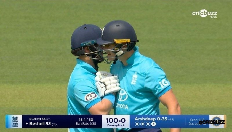 Two cricket players in light blue uniforms and navy helmets lean together on a grass field during a live broadcast, with a scorecard reading "100-0."