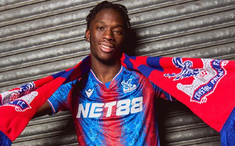 A young man poses wearing a blue and red Crystal Palace jersey with "NET88" printed on it, holding a matching scarf behind his neck.