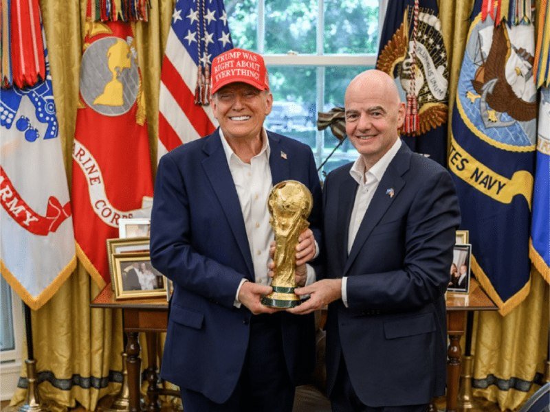 Two men in suits pose in an ornate room holding a gold World Cup trophy, standing before American and military flags, with one man wearing a red "Trump Was Right About Everything" cap.