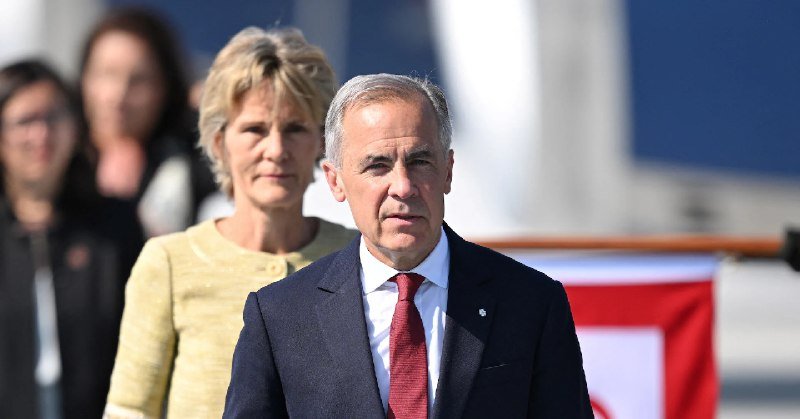 A man in a dark suit and red tie stands in the foreground, with a woman in a yellow dress behind him and other figures blurred in the background.