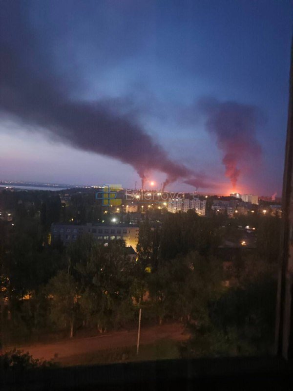 Plumes of dark smoke rise from distant fires in a city skyline at dusk, viewed through a window above silhouetted trees and buildings.