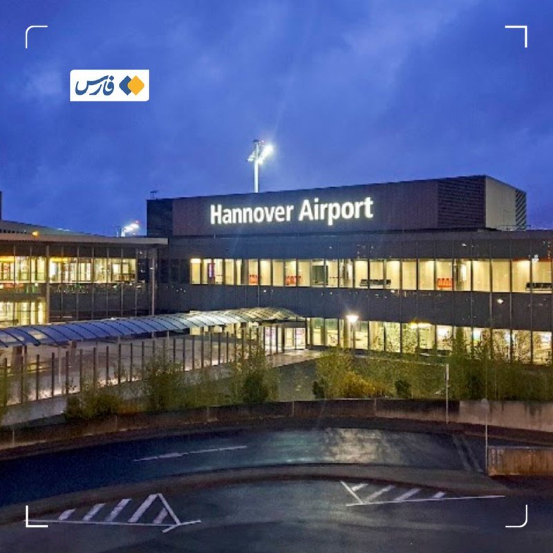 The illuminated terminal building of "Hannover Airport" stands against a dusk sky, with lit interior windows and an empty foreground roadway visible.