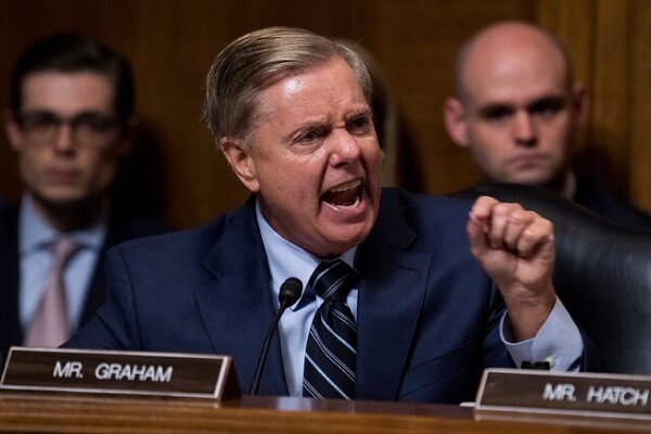 A man in a dark suit gestures emphatically while speaking at a wooden desk with a "MR. GRAHAM" nameplate, with two other men seated behind him.
