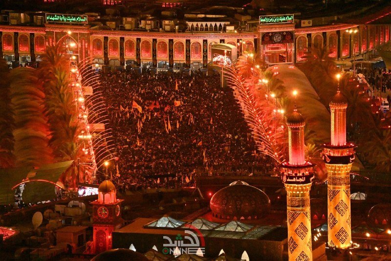 Aerial nighttime view of a vast illuminated shrine courtyard filled with crowds, flanked by golden minarets and palm trees under glowing red arches.