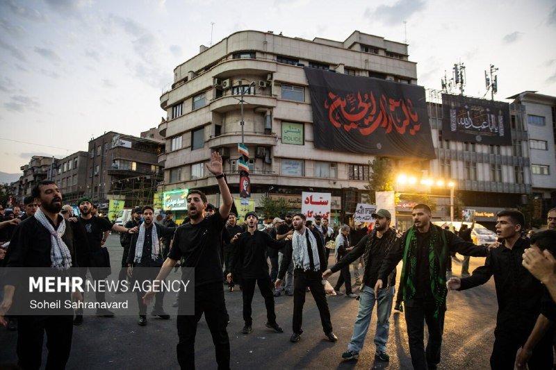 A crowd waves Iranian flags and holds up framed portraits during a public gathering outside a multi-story building under construction, with logos overlaid in the upper corner.