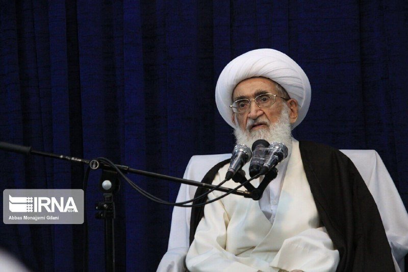 An elderly cleric with a white beard, glasses, and white turban speaks into a microphone, gesturing with his ringed hand, bearing a "TASNNIM NEWS" watermark.