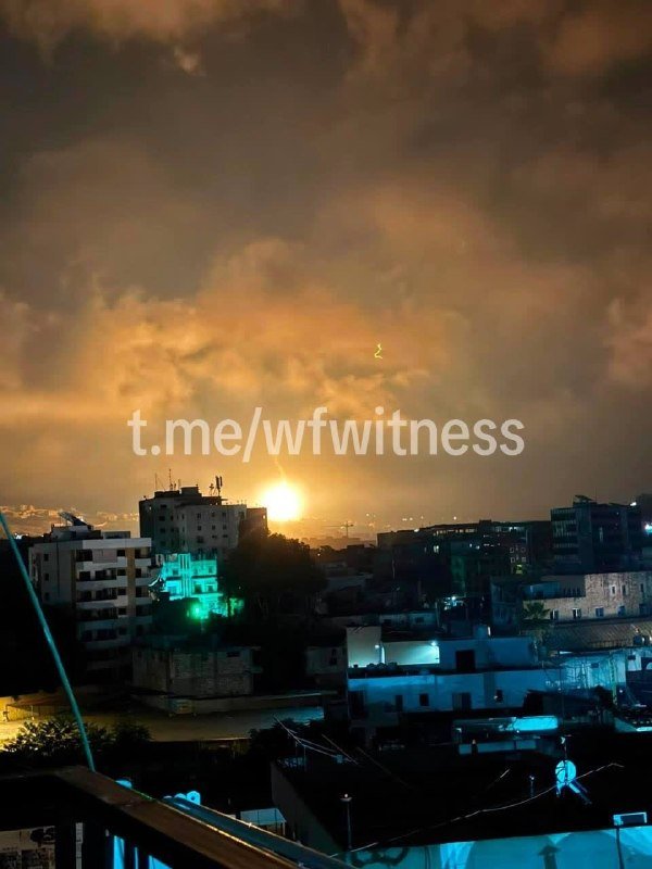 A bright light rises over a nighttime cityscape of densely packed buildings beneath cloudy skies, with a "t.me/wfwitness" watermark across the center.