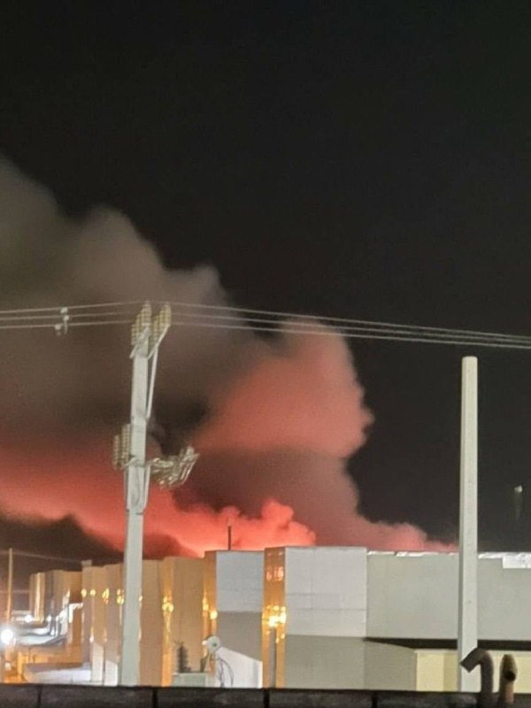 Plumes of smoke illuminated in red rise above industrial buildings and a utility pole at night.