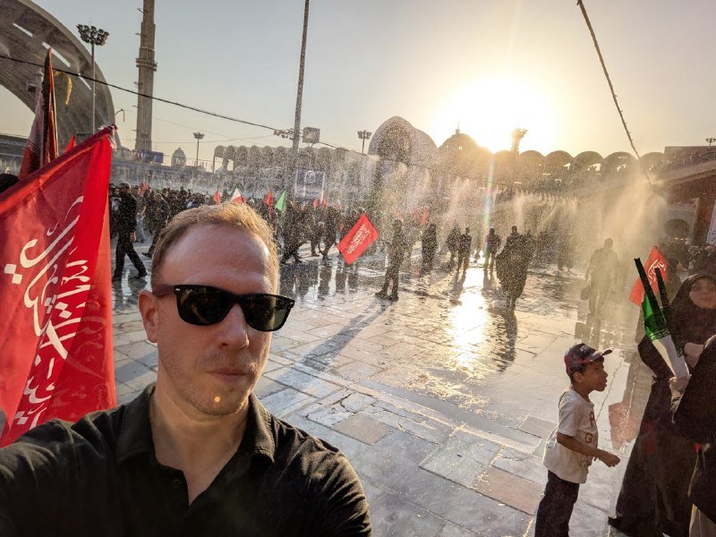 A crowd of people, many holding red flags and a portrait of a bearded man in religious attire, gather in a city street during a daytime demonstration.