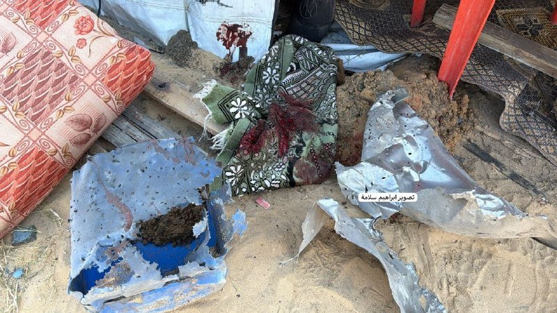 Debris including torn patterned fabric, a blood-stained rug, sand-covered items, and a red metal structure lie scattered on sandy ground.