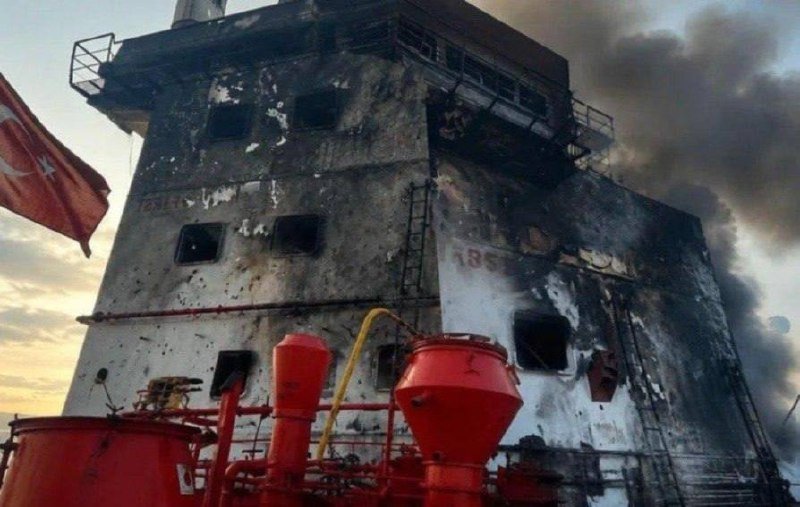 A burnt, multi-story building structure is visible with blackened walls, open window frames, and dark smoke rising nearby.