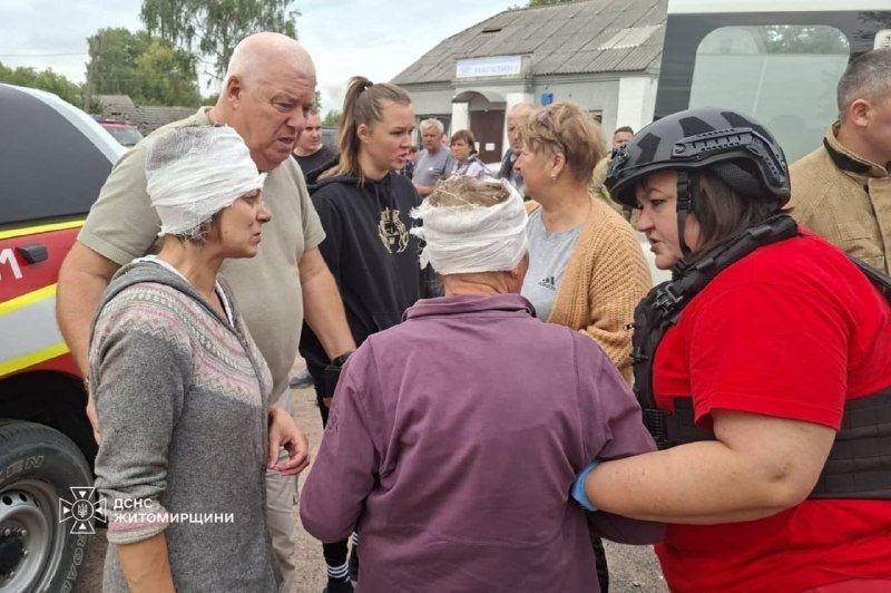 A group of civilians, some with bandaged heads, stand outdoors being assisted by a rescue worker in a red uniform and helmet, with emergency vehicles visible in the background.