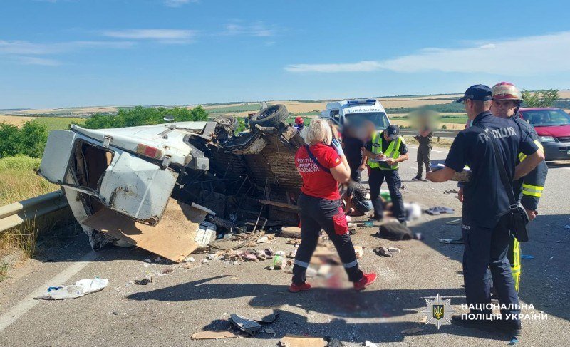 An overturned vehicle lies on a rural road as emergency responders in high-visibility vests and uniforms inspect the crash scene under a clear blue sky.