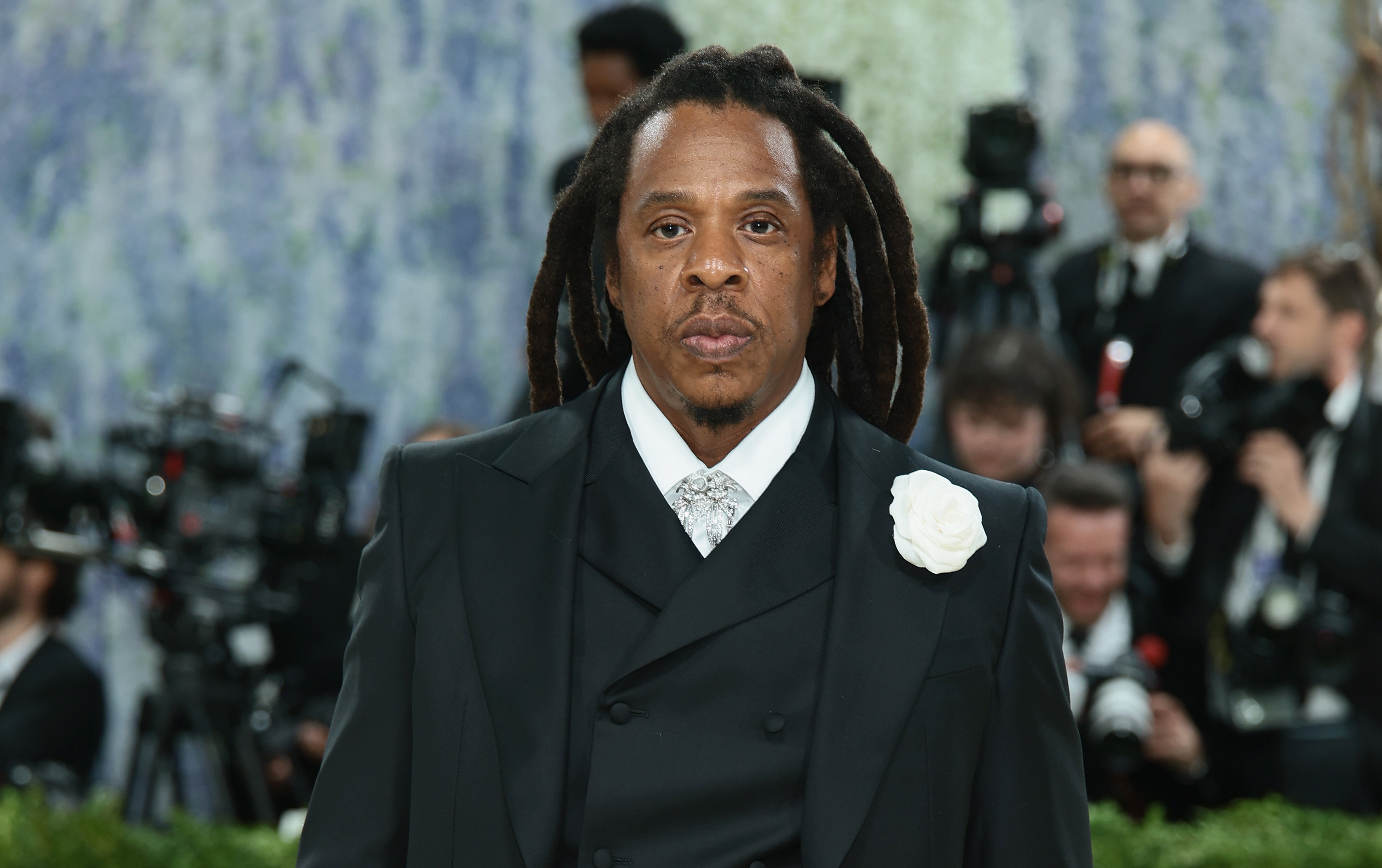 A man with long dreadlocks wearing a black double-breasted suit and white rose boutonnière poses before photographers and cameras at a formal event.