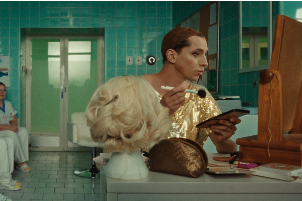 A person in a gold sequined outfit applies makeup to a blonde wig on a mannequin head in a tiled room, while another person sits nearby.