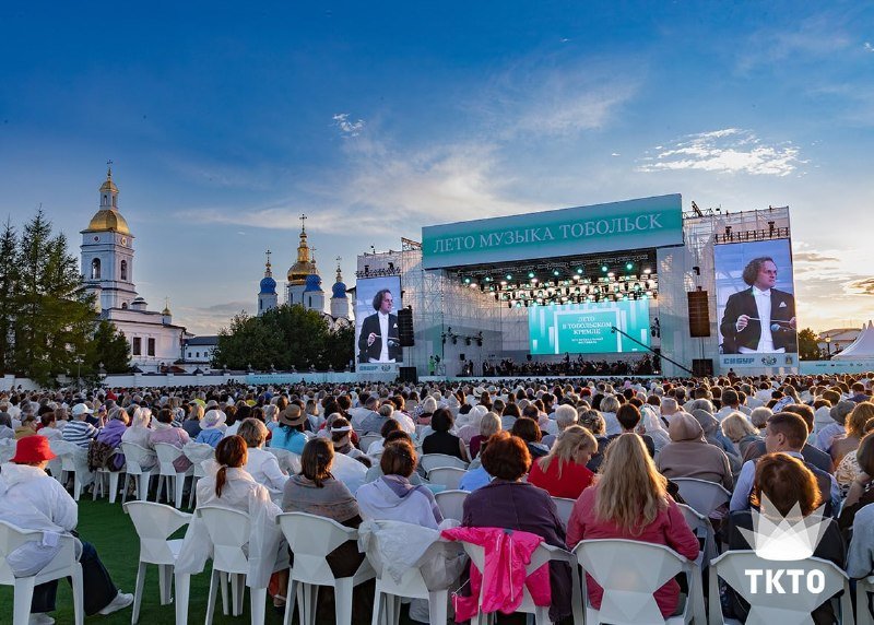 A large outdoor audience sits in white chairs facing a concert stage with screens displaying "ЛЕТО МУЗЫКА ТОБОЛЬСК," beside a Russian Orthodox church complex with golden and blue domes.
