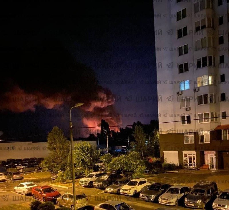 A nighttime view shows a parking lot filled with cars beside a tall residential building, while thick smoke and a reddish glow rise over the distant treeline.