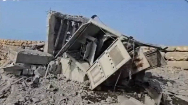 A collapsed concrete and steel structure lies in rubble under a clear blue sky, with sandbags stacked in the background.
