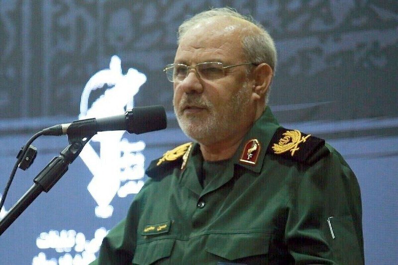 An older bearded man in a green military uniform with insignia stands at a microphone, appearing to speak at a podium against a dark backdrop.