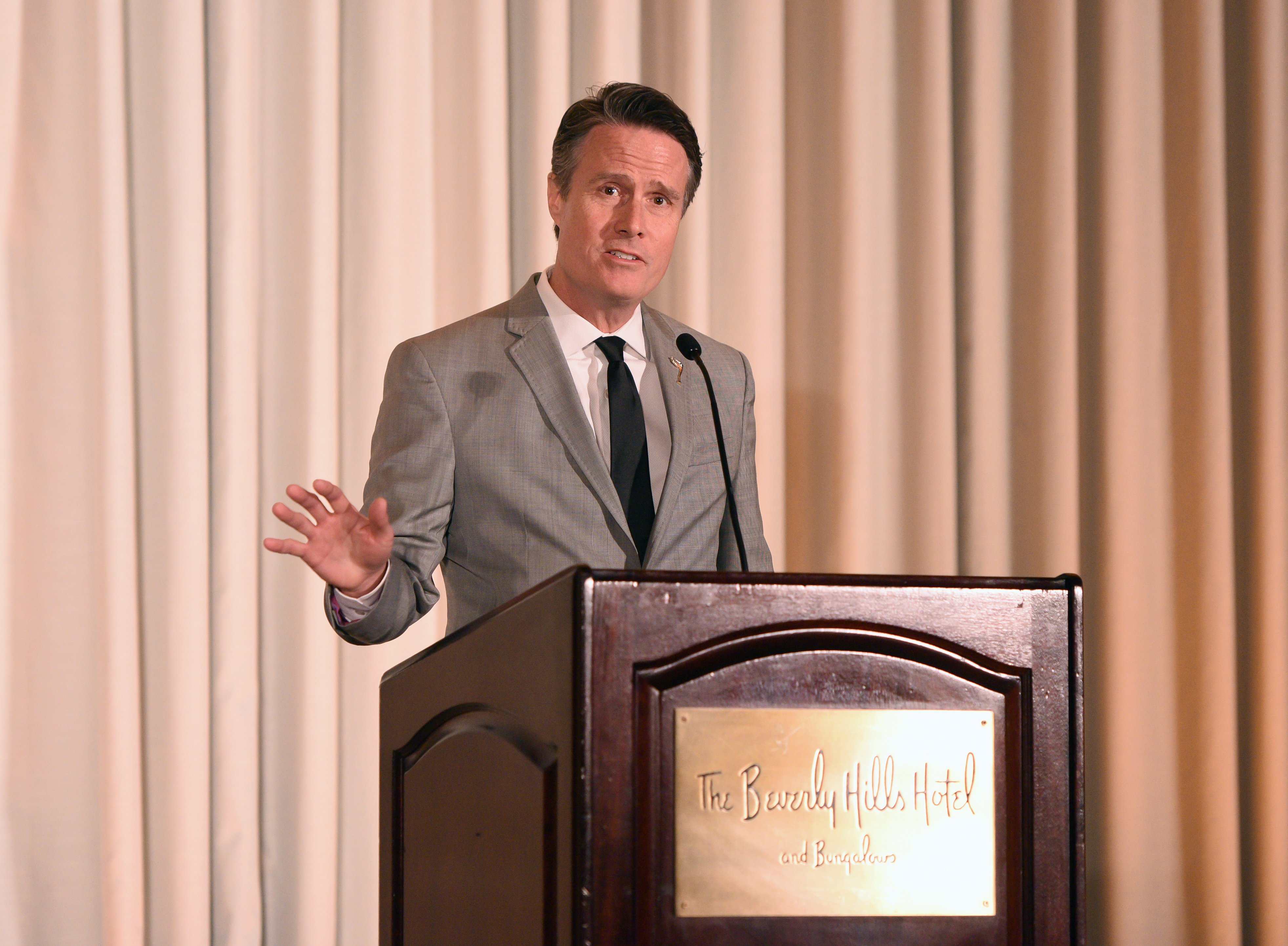 A man in a gray suit and black tie speaks at a wooden podium bearing a "The Beverly Hills Hotel and Bungalows" plaque, gesturing with one hand in front of cream-colored curtains.
