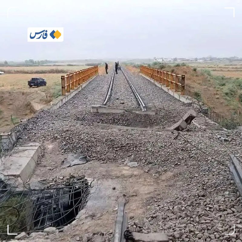 A damaged railway bridge with broken tracks, exposed rebar, and scattered ballast is shown with two people standing mid-span and a vehicle parked on an adjacent dirt road.