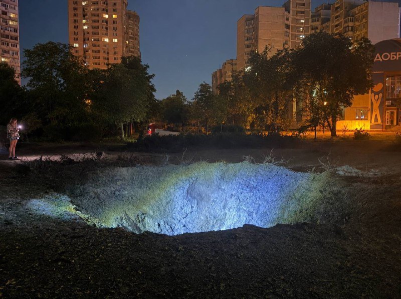 A large crater in the ground is illuminated with blue and green light at night, surrounded by trees and high-rise apartment buildings, with a person holding a flashlight nearby.