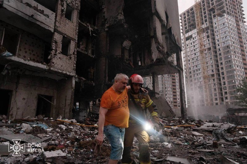 A firefighter in a red helmet assists a man in an orange shirt through rubble beside a heavily damaged building, with a high-rise under construction in the background.